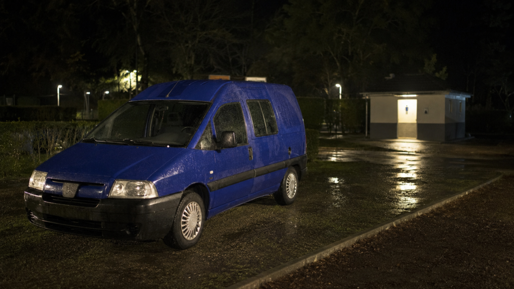 Furgoneta camper aparcada de noche bajo la lluvia junto a los baños de un camping.