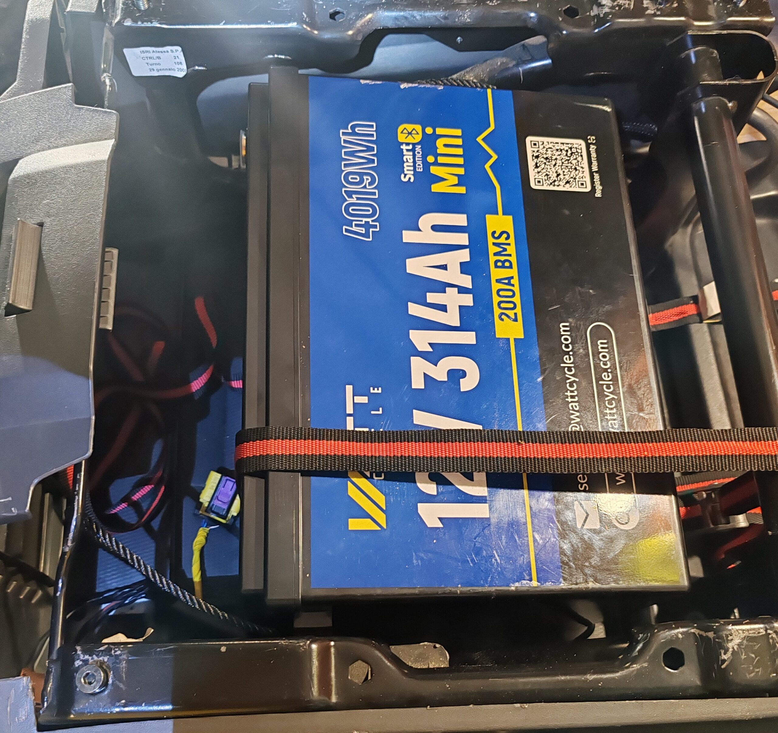 Batería LiFePO₄ WattCycle de 314 Ah sujeta con cincha bajo el asiento del conductor de la autocaravana.
